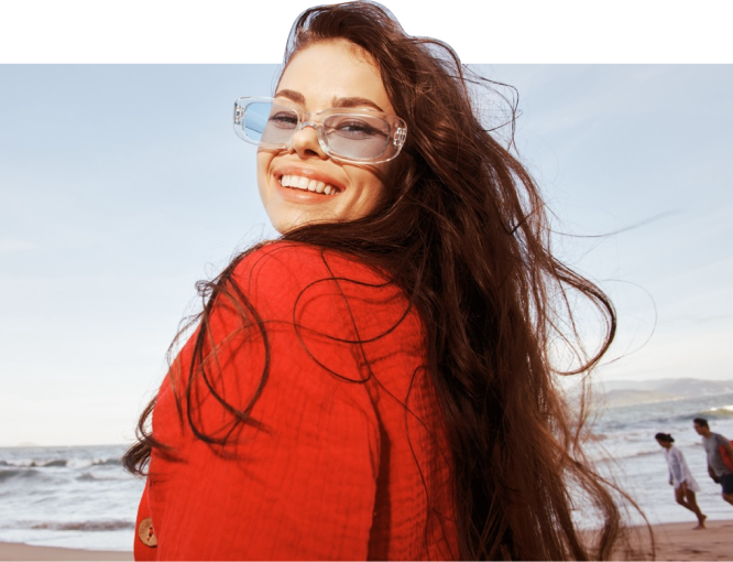 Happy woman in a red jacket and sunglasses at a beach, with waves behind her.